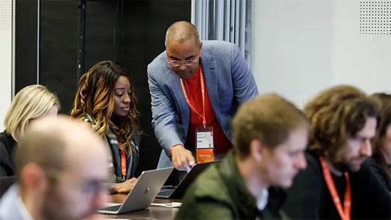 Group of people in a room with a man helping show a woman something on a computer.